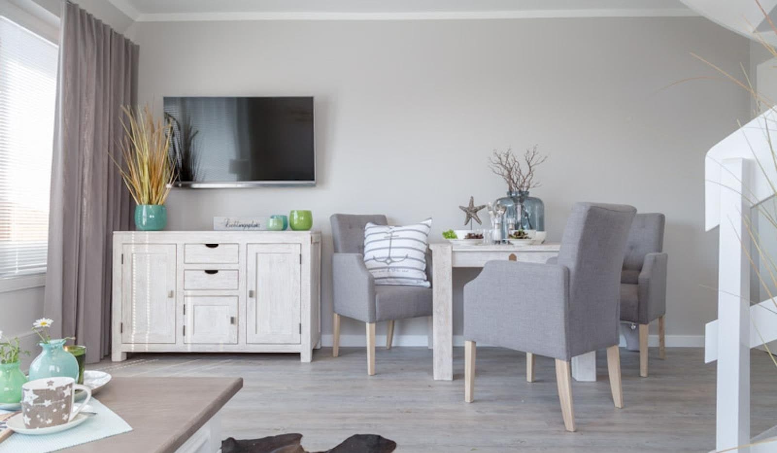 Wohnzimmer mit Essplatz, drei grau gepolsterten Stühlen, Sideboard in Weißholz und Flachbildfernseher an der Wand